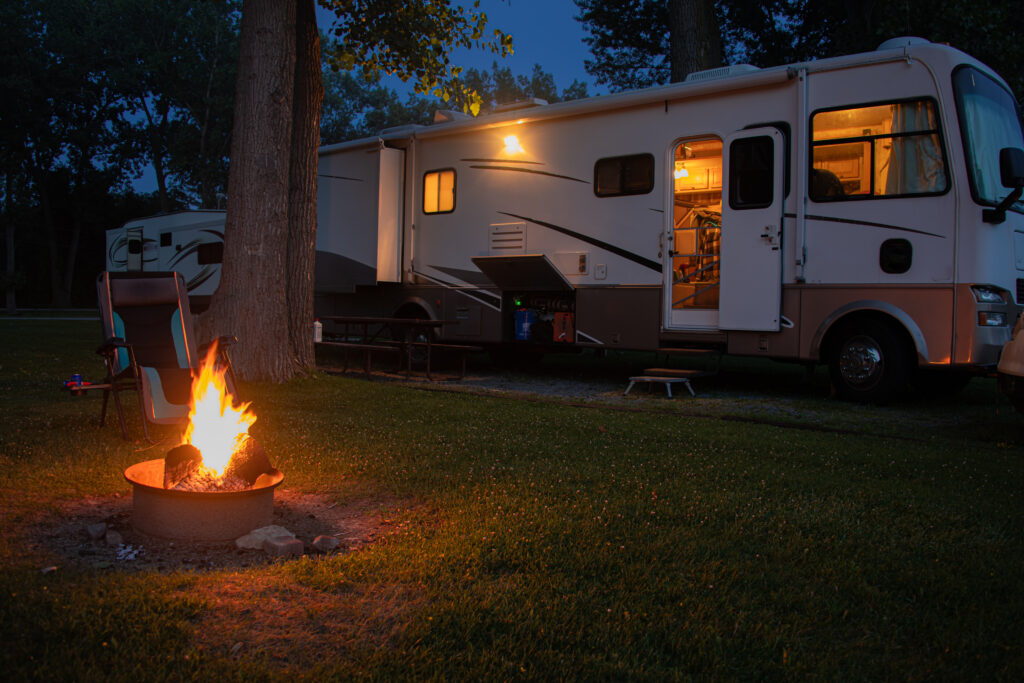 RV motorhome parked at a site at night with its inside lights on and a small campfire outside